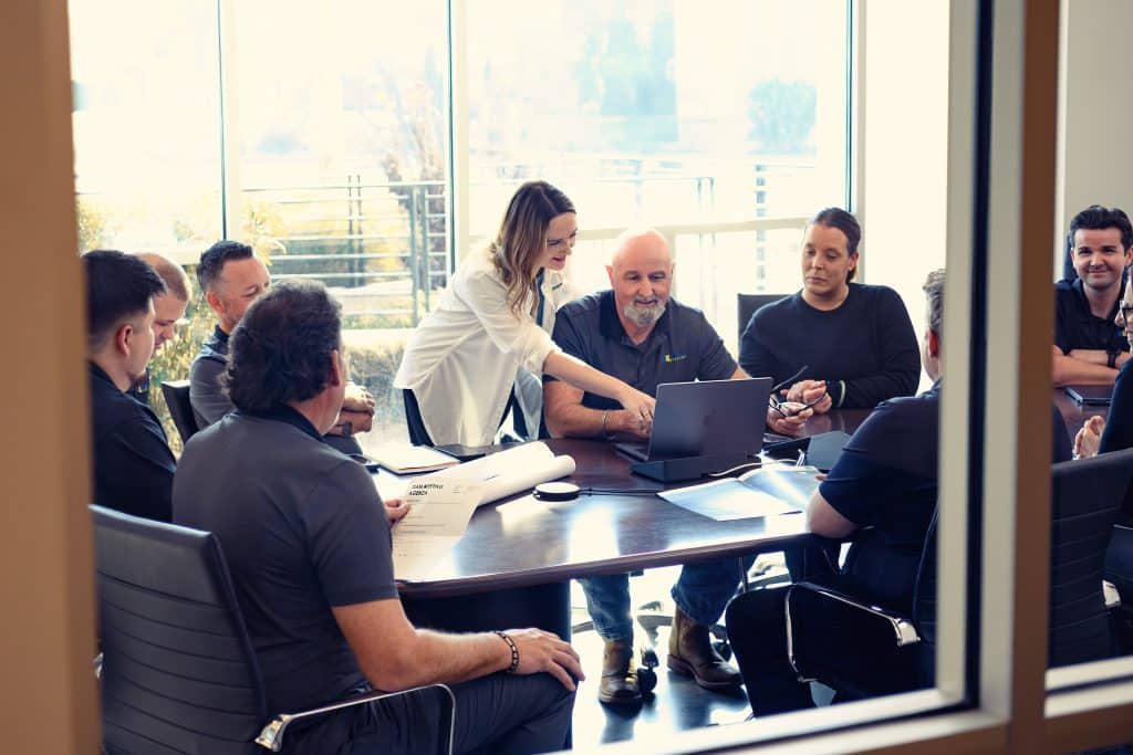 Eight people gathered around a table in an office, discussing something on a laptop.