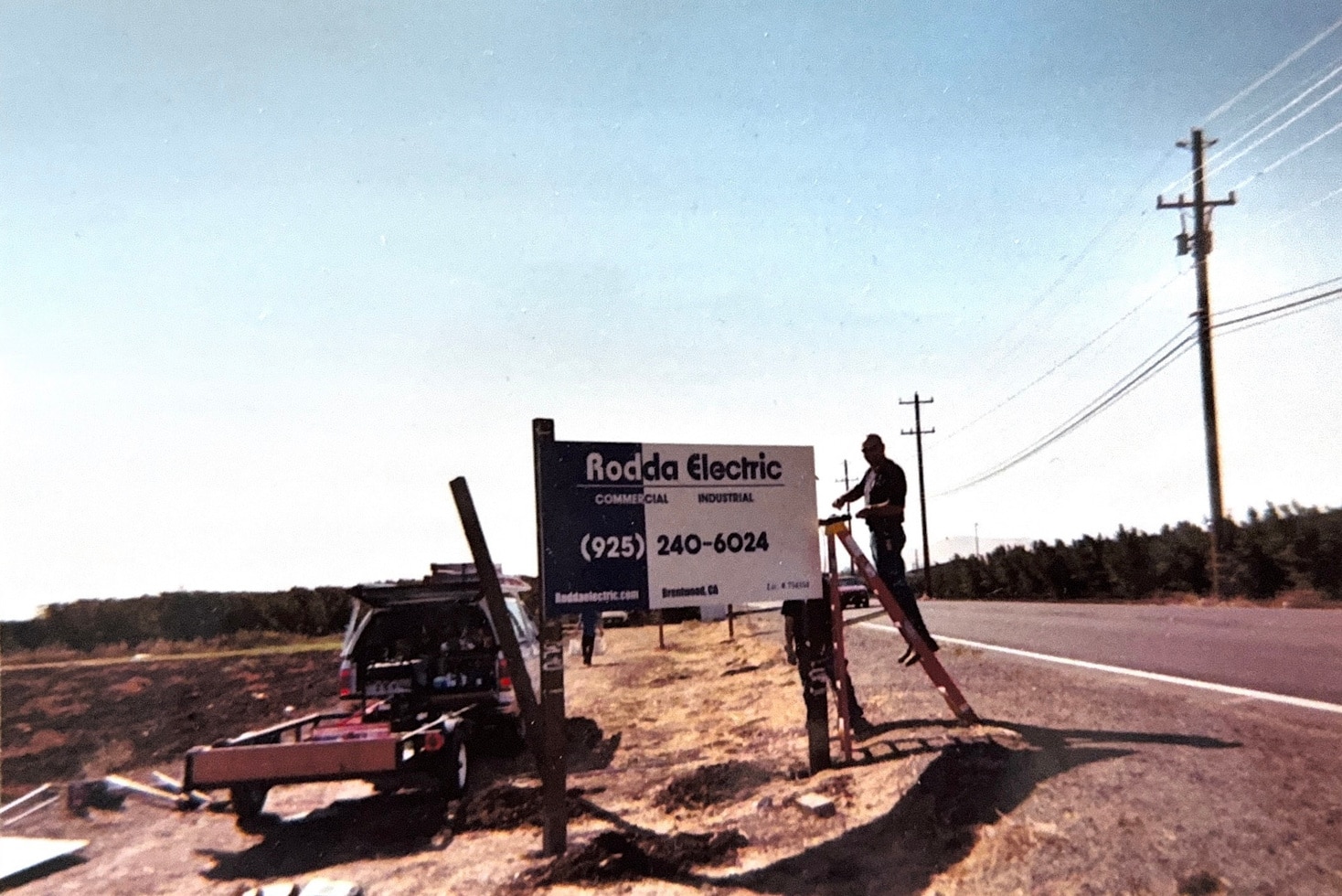 Person on ladder installs Rodda Electric sign by a roadside with utility poles nearby.