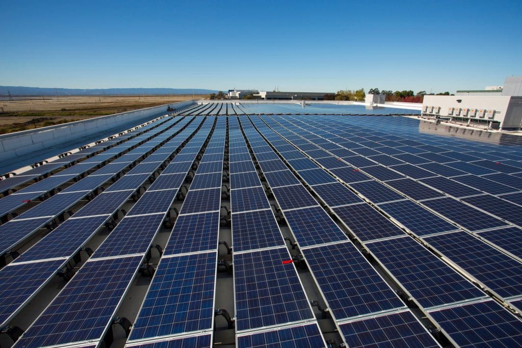 Rows of solar panels installed on a large, flat rooftop under a clear blue sky.