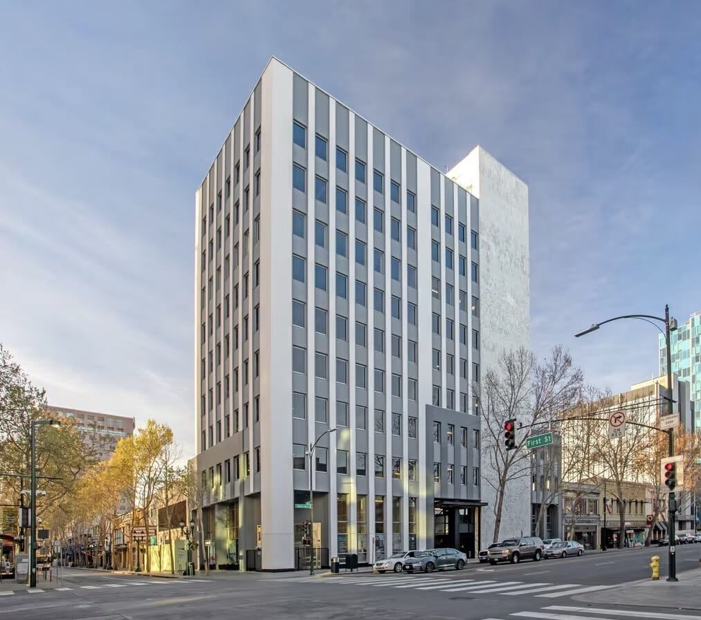 A modern, mid-rise office building on a city street corner with cars and trees nearby.