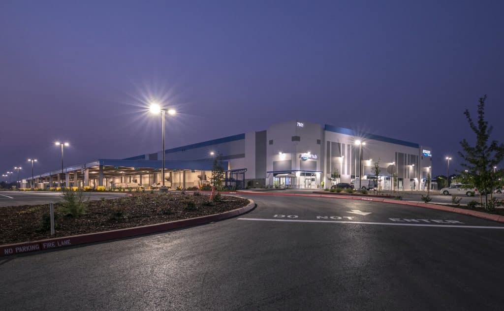 Large warehouse building with outdoor lights on, surrounded by parked cars at dusk.