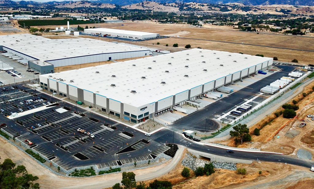 Aerial view of a large warehouse with parking lots and trucks, surrounded by open land.