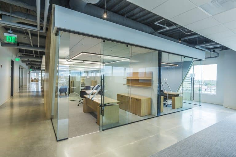 Modern glass-walled office with desks, chairs, and shelves in a bright, open hallway.