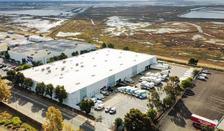 Aerial view of a large warehouse with trucks and wetland area in the background.