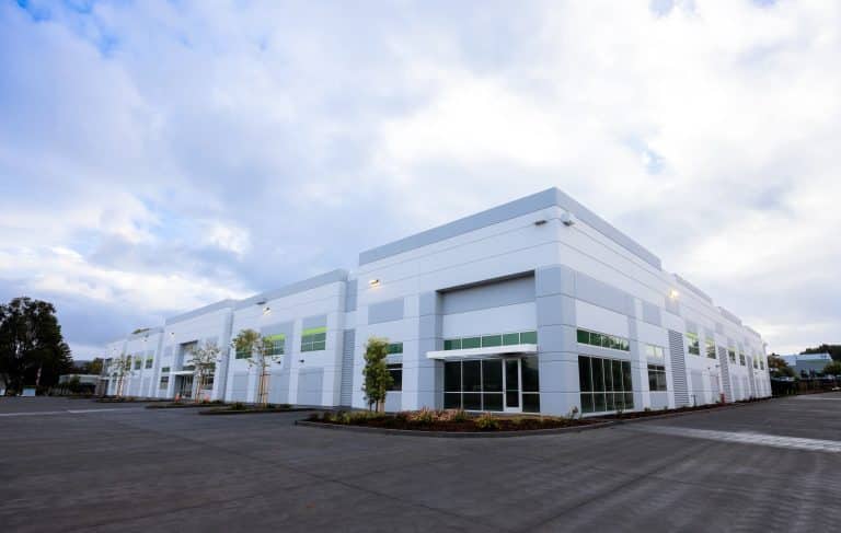 Modern white industrial building with large windows and an empty parking lot outside.
