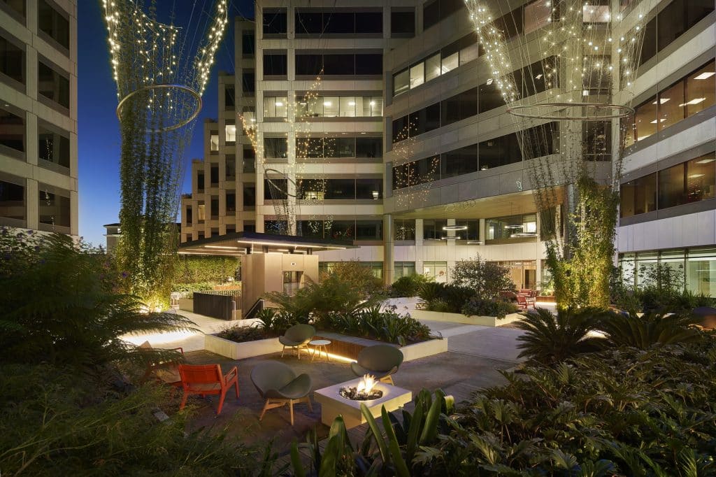 Modern courtyard at night with string lights, plants, and seating around a fire pit.