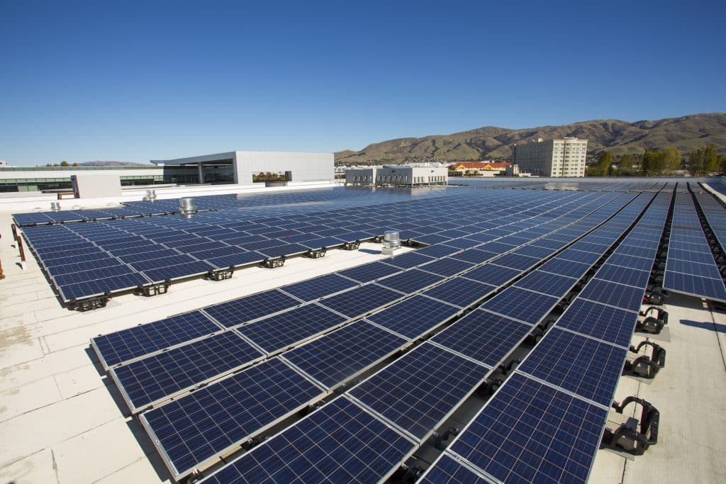 Rows of solar panels on a rooftop under a clear blue sky with mountains in the background.