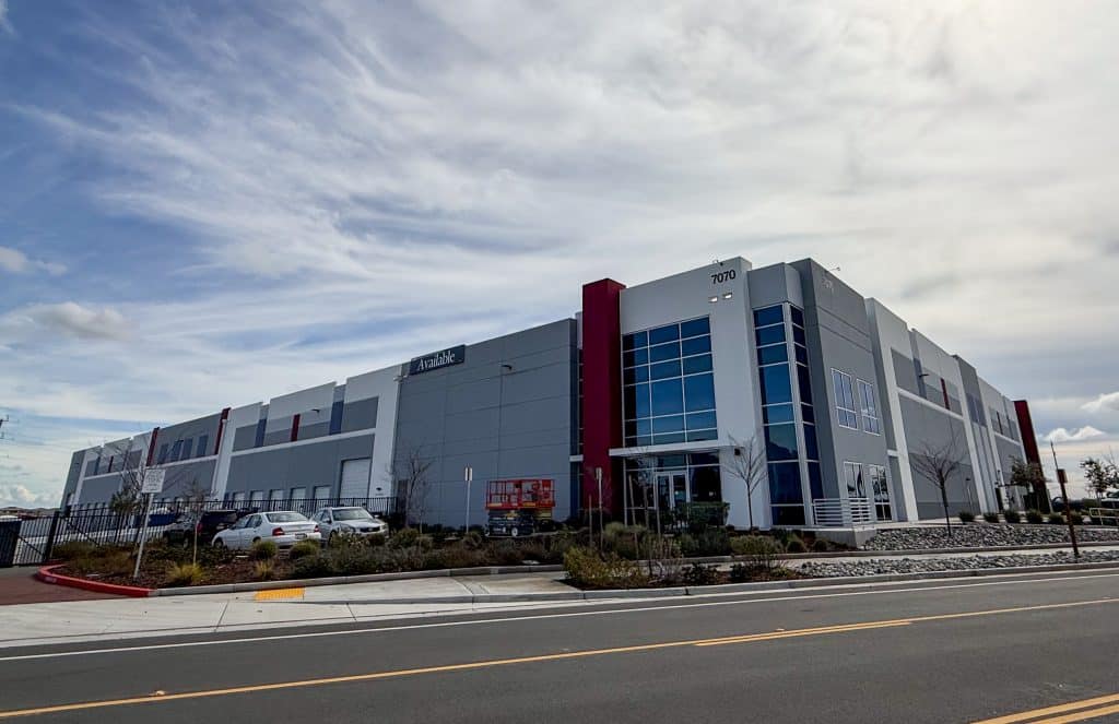 Modern warehouse building with large windows, parked cars, and a cloudy sky above.