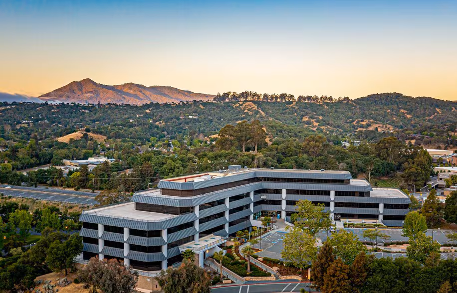 Modern office building surrounded by trees with hills and mountains in the background.