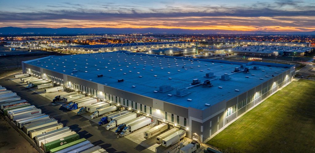 A large warehouse with many trucks parked outside at sunset in an industrial area.