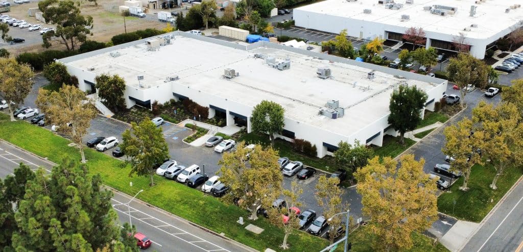 Aerial view of a white commercial building with cars parked around and trees nearby.