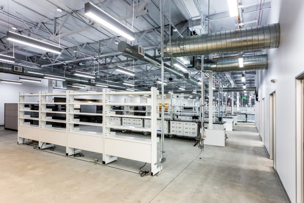 Modern, empty laboratory with white workstations, shelves, and exposed ductwork.