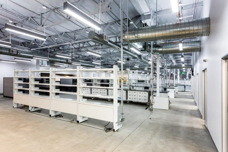 Modern, empty laboratory with white workstations, shelves, and exposed ductwork.