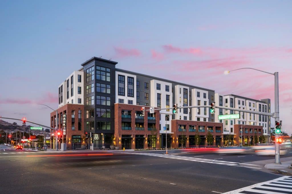 Modern, multi-story apartment building at a busy intersection during sunset.