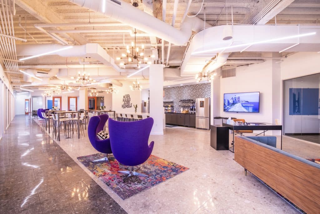 Modern open office space with purple chairs, high tables, and a kitchen area.