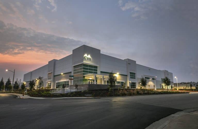 Modern white warehouse building at sunset with lights on and a landscaped exterior.