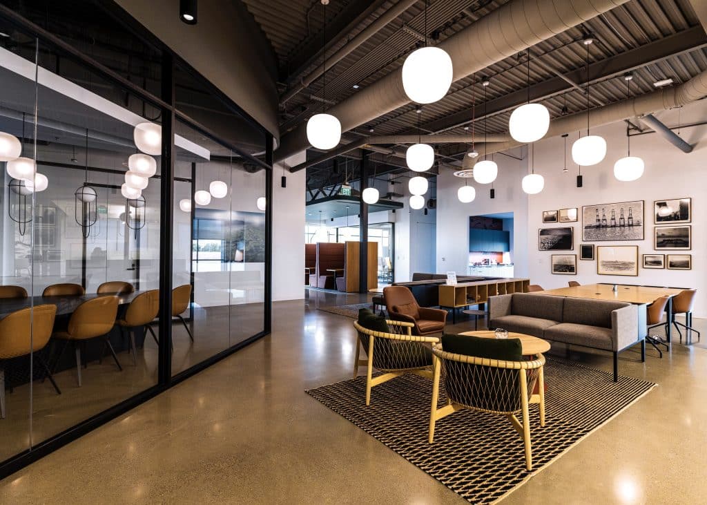 Modern office lounge with sofas, chairs, hanging lights, and a glass-walled meeting room.