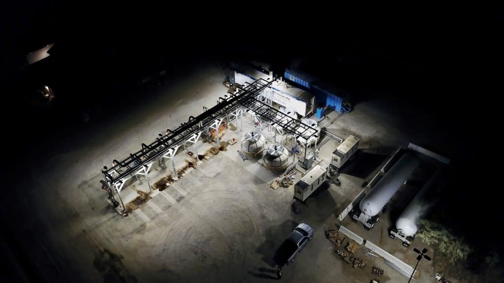 An industrial facility at night, brightly lit, with tanks, pipes, and parked vehicles visible.