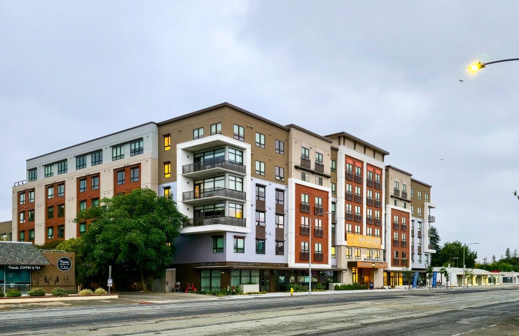 Modern mid-rise apartment building with balconies, shops below, and cloudy sky above.