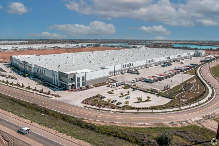 Aerial view of a large white warehouse with loading docks, trucks, and parking areas.