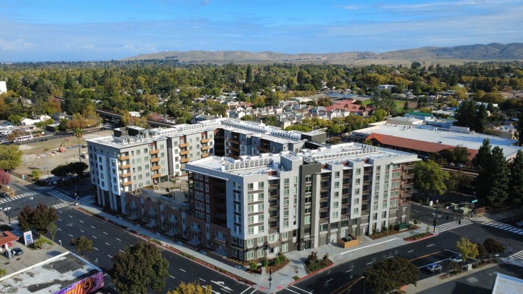 A modern mid-rise apartment building in a suburban area with trees and hills in the background.