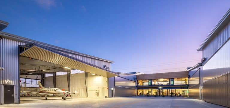 Small airplane in a modern hangar at sunset with glass-walled building in the background.