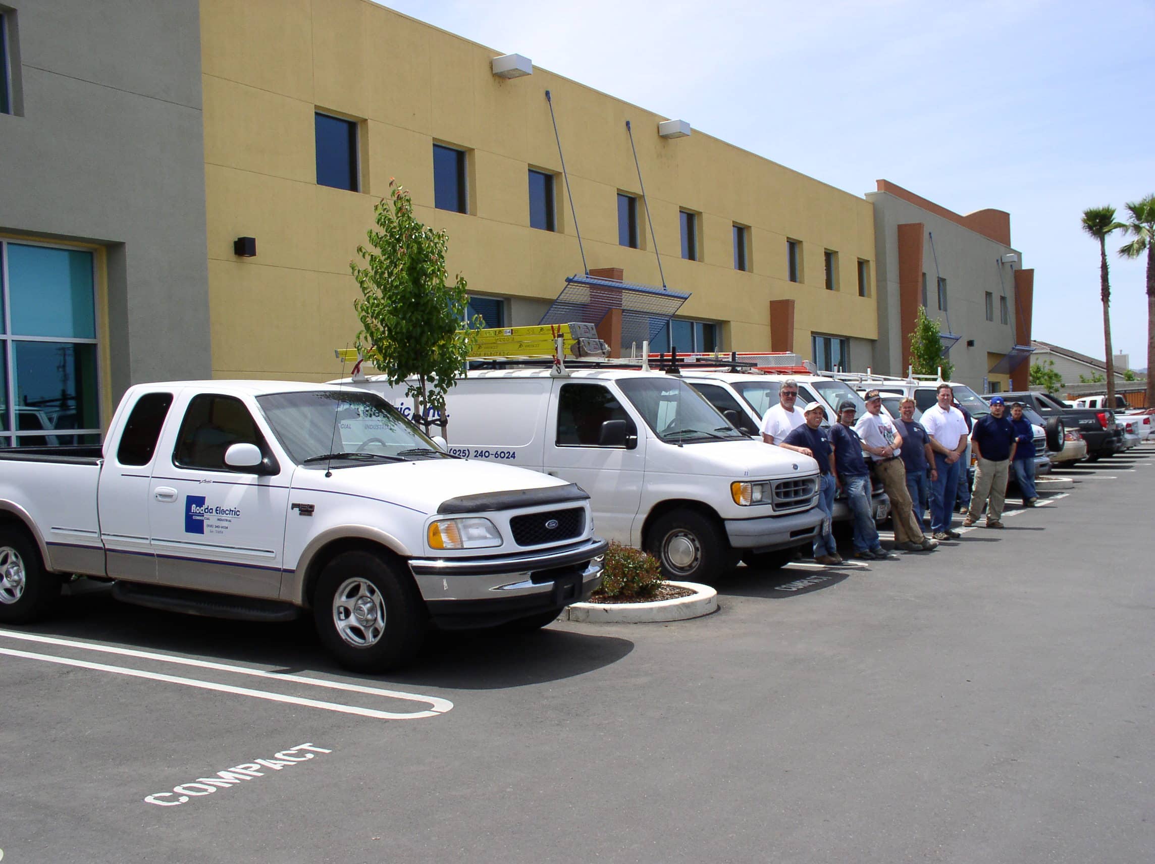 People stand by work trucks and vans parked outside a modern commercial building.